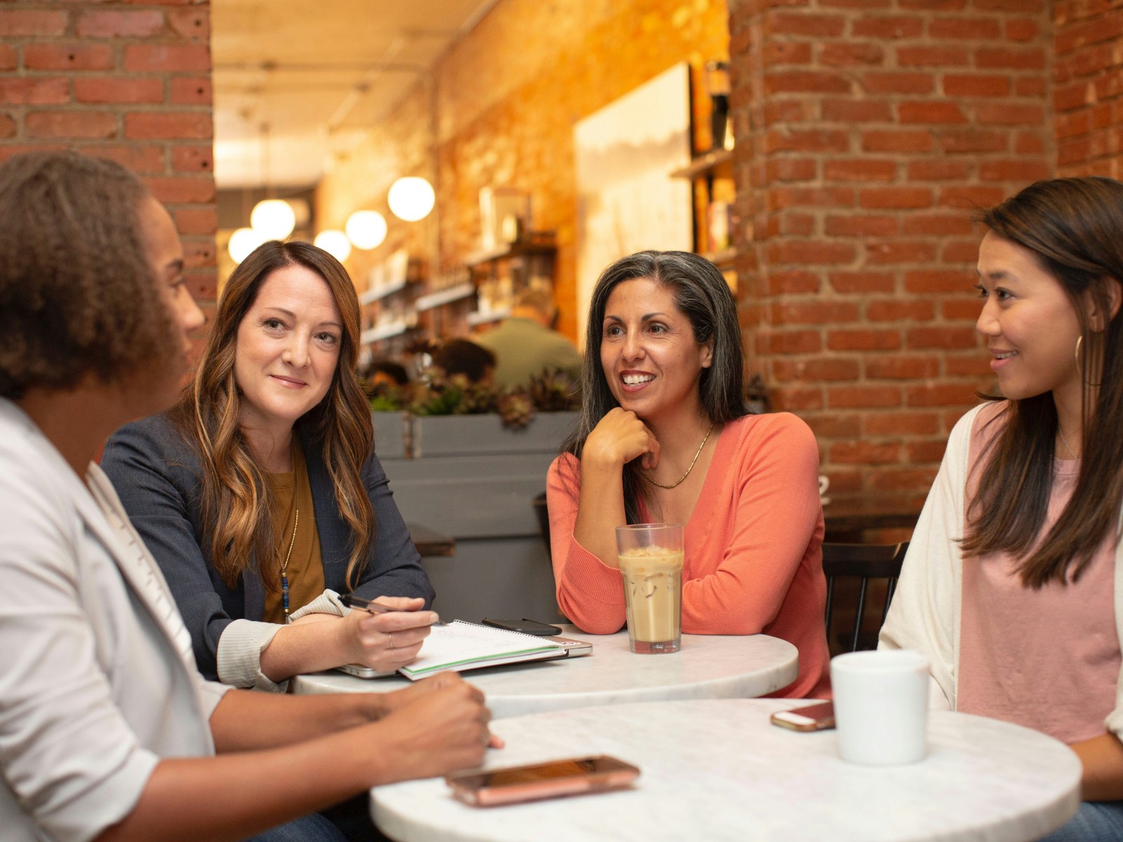 Four people in conversation around a café table, notebook open.